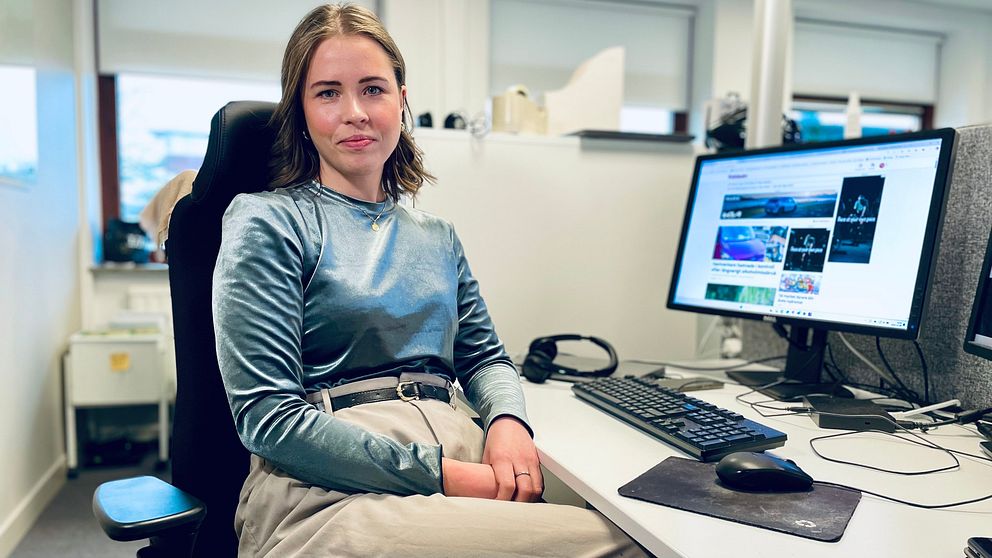 En person sitter vid en arbetsstation med en dator framför sig. Bakgrunden visar en kontorsmiljö med ljusa fönster. Datorn visar en webbplats, och det finns en mus och ett tangentbord på skrivbordet. Personen bär en långärmad tröja och byxor, och sitter avslappnat i en ergonomisk stol.