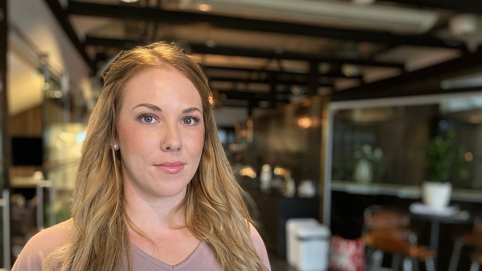 En person med långt, vågigt hår står i en modern inomhusmiljö. I bakgrunden syns eleganta möbler och växtlighet.
