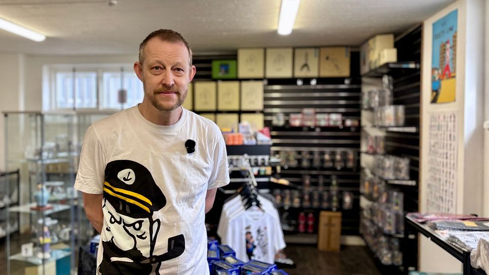 Person med vit t-shirt står i butik med hyllor fyllda av olika varor och tryckta t-shirts.