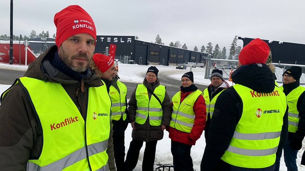 Flera personer med gula västar där det står ”konflikt” i röd text står framför Teslas svarta anläggning.