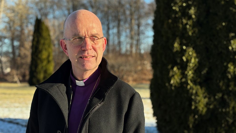 Biskop Fredrik Modéus iklädd vinterrock står utomhus i vintrig park med barrträd i bakgrunden.