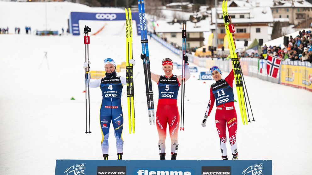 Tre skidåkare står på prispallen med sina skidor i luften, glada över sina prestationer. Den första åkaren, klädd i en röd tröja med nummer 5, står i mitten och håller upp sina skidor. De två andra åkarna, en i blå och en i svart outfit med nummer 4 och 11, står på vardera sida om henne. I bakgrunden syns snötäckta fjäll och en stor publik.