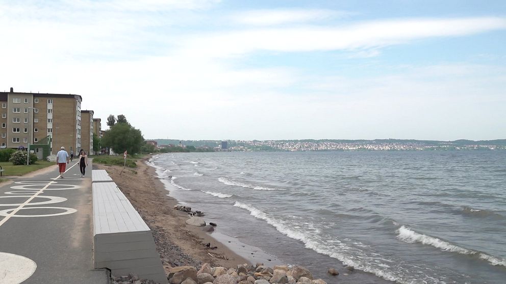 Vätterstranden i Jönköping. Till vänster skymtas hus i bakgrunden, på gångvägen promenerar två personer. Närmast strandkanten finns bänkar att sitta på.