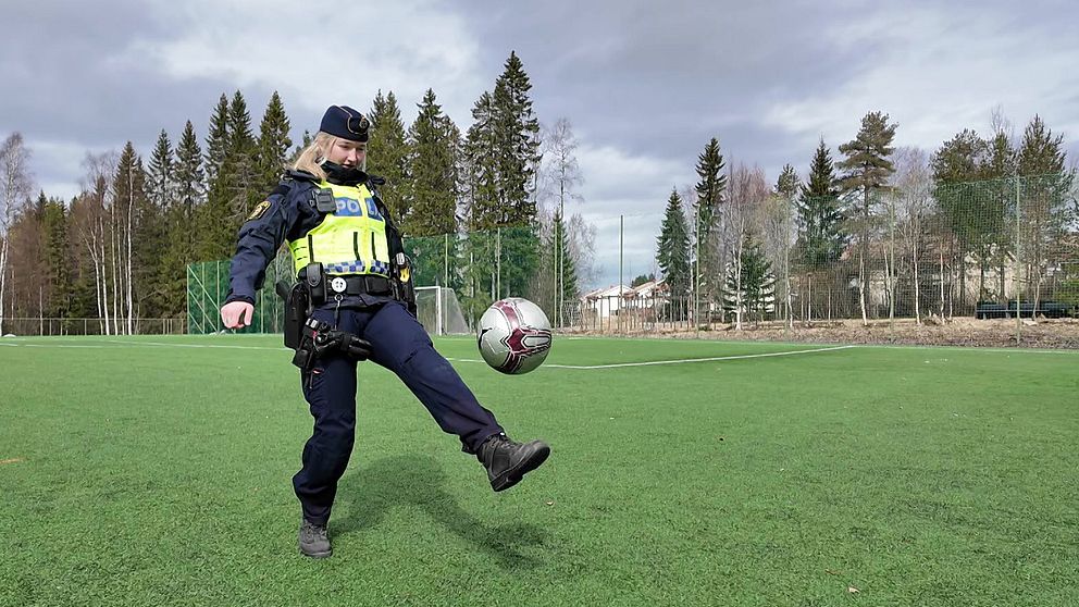 En polis jonglerar med en fotboll på en gräsplan utomhus framför träd och skog.
