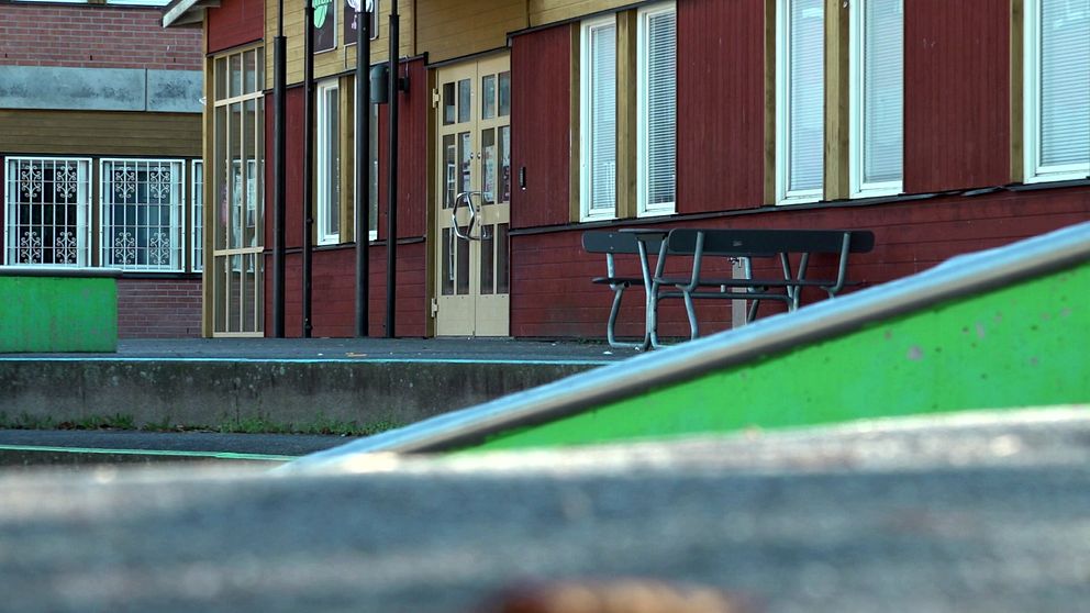 En delad bild som visar en del av en skolbyggnad. I förgrunden syns en grön ramp och en betongyta. I bakgrunden är skolans huvudentré med fönster och dörrar, samt en bänk som står vid väggen. Det finns också gröna och röda färgdetaljer på byggnaden.