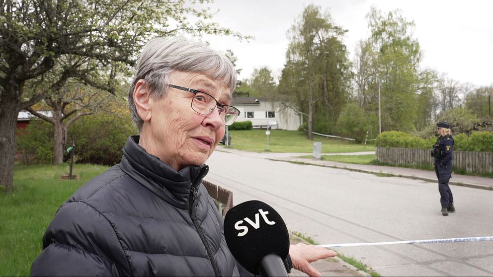 Reporter håller SVT-mikrofon mot en äldre kvinna, polis står bakom avspärrning på lugn villagata.