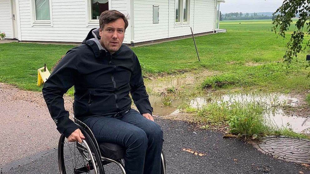 Man i rullstol utomhus framför vitt hus och en vattenpöl