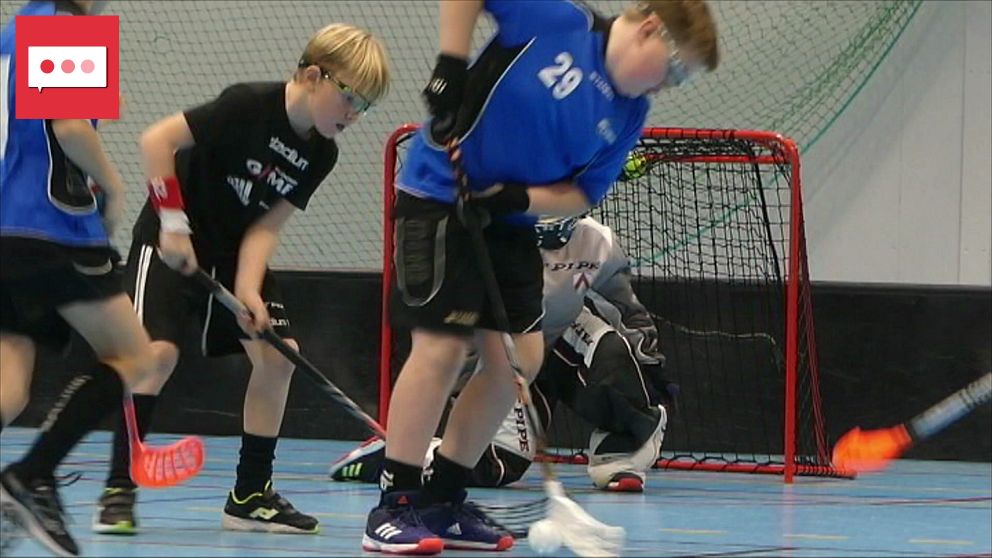 Tre unga spelare spelar innebandy på en sporthall, med en målvakt i bakgrunden.