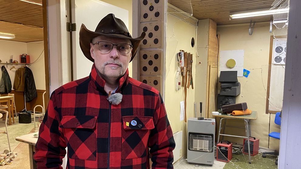 Person i röd- och svartmönstrad skjorta, cowboyhatt, står i verkstad med verktyg på vägg.