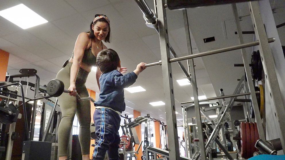 Mamma tittar på sitt barn på gymmet och håller en vikt, ser glad ut