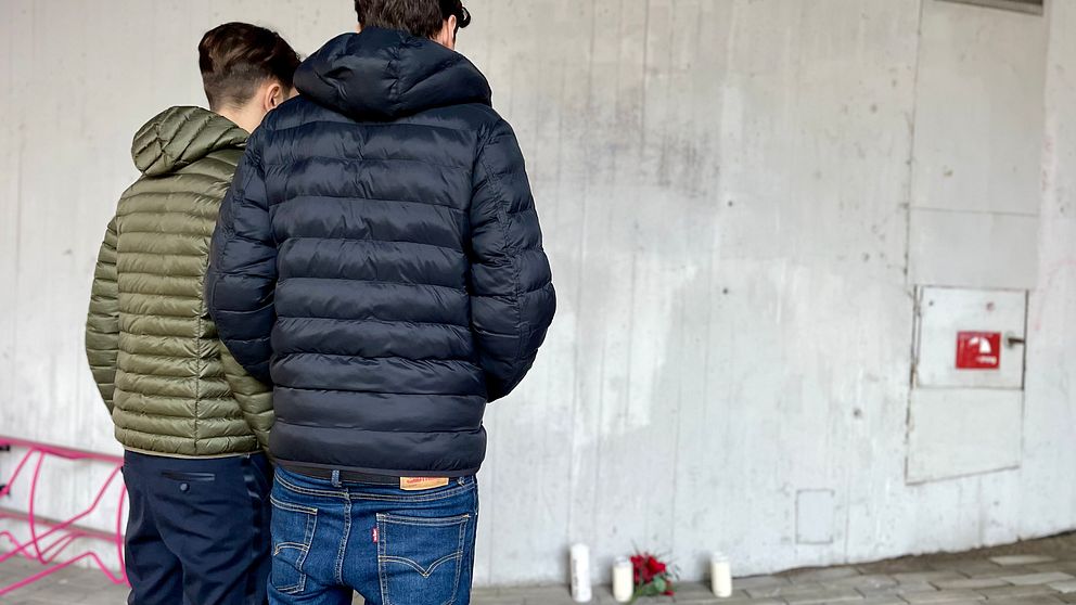 Två personer i dunjackor står med ryggen mot kameran, de betraktar ett blommor och ljus mot en betongvägg. Bakgrunden är enkel och förstärker den stillsamma stämningen.