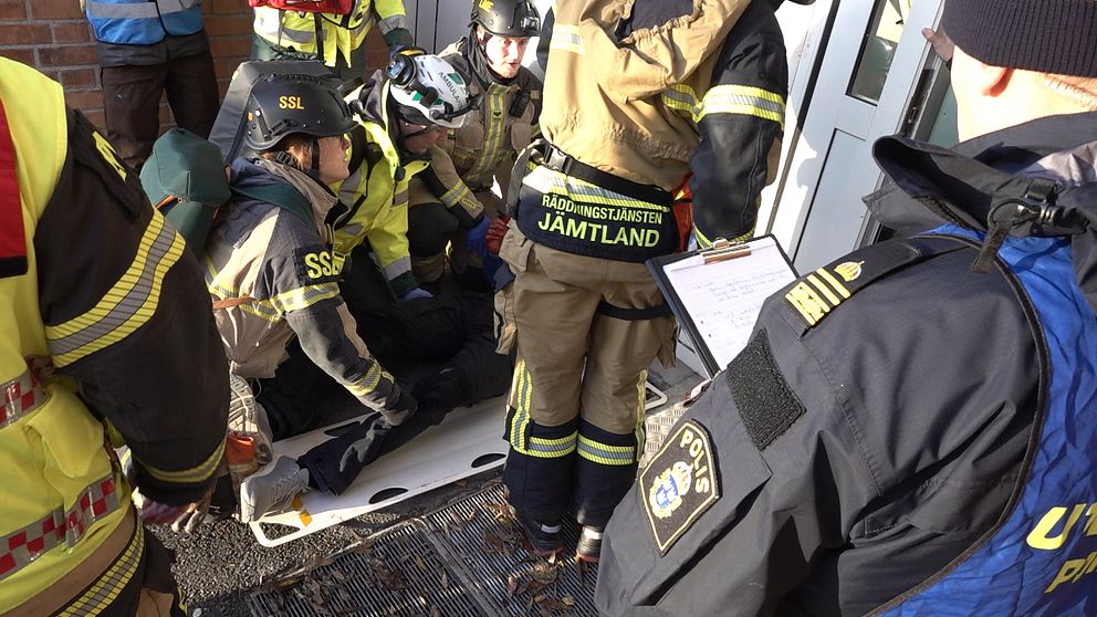 Brandmän och polis arbetar tillsammans för att hjälpa en skadad person som ligger på en bår utanför en byggnad. Scenen visar en samordnad insats av räddningstjänsten och polisen.