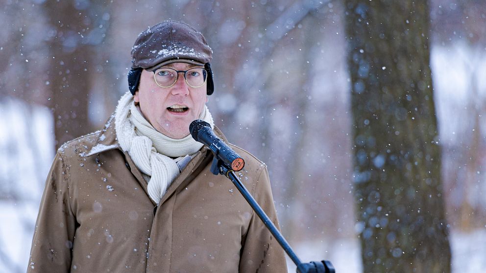 En person klädd i en brun jacka och vit scarf står framför en mikrofon i en snöig miljö.