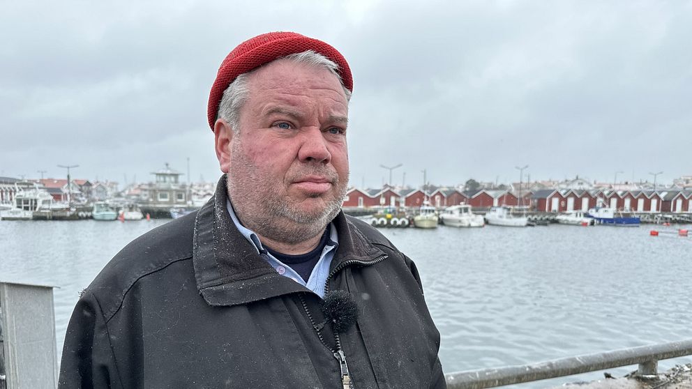 Skäggig man med svart jacka och röd mössa står vid hamn med röda sjöbodar och båtar i bakgrunden.