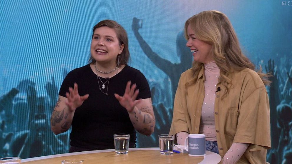 Två personer sitter vid ett bord i en tv-studio, bakom dem skymtar en konsertpublik.