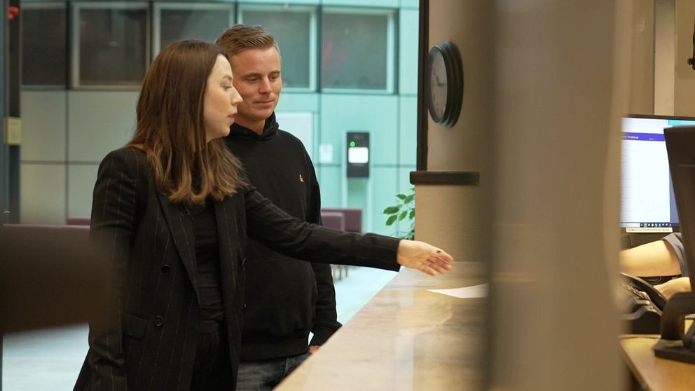 Jonathan Ravelin och Elsa Häggård står vid en receptionsdisk och lämnar över ett dokument. Bakgrunden visar en modern kontorsmiljö med växter och en klocka på väggen. Skärmen på disken är delvis synlig.