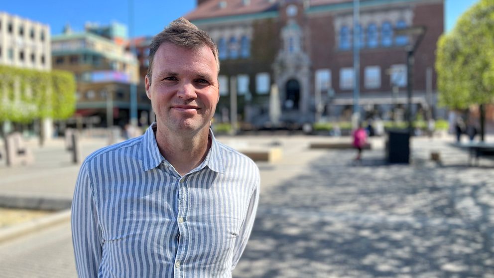 Person i randig skjorta står på soligt torg, byggnad syns i bakgrunden.