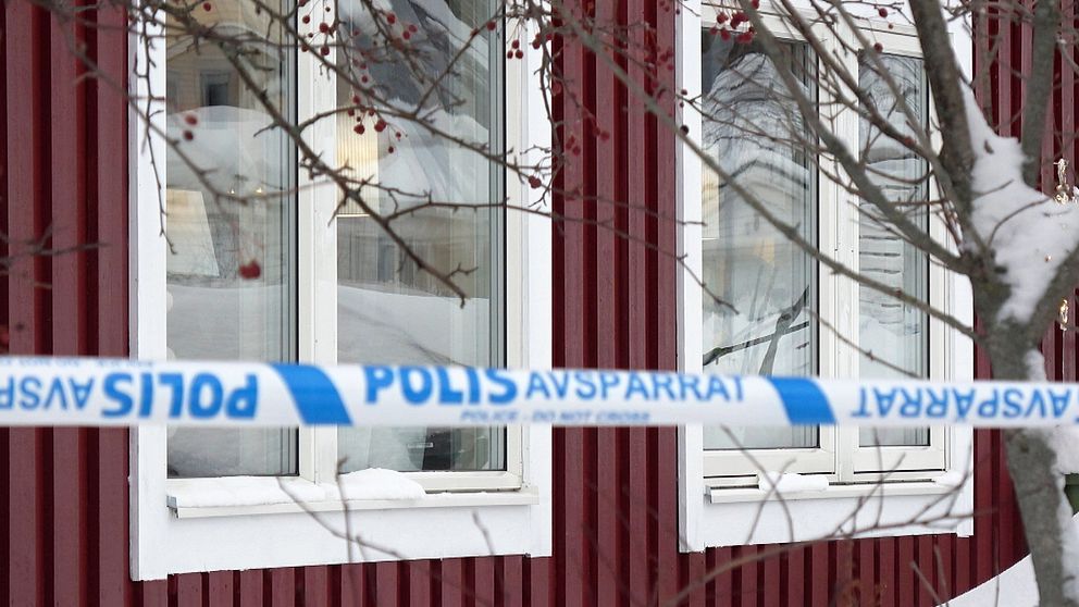 Fönster på ett rödbrunt hus inhägnat med blåvita polisavspärrningsband och grenar från ett träd i förgrunden.