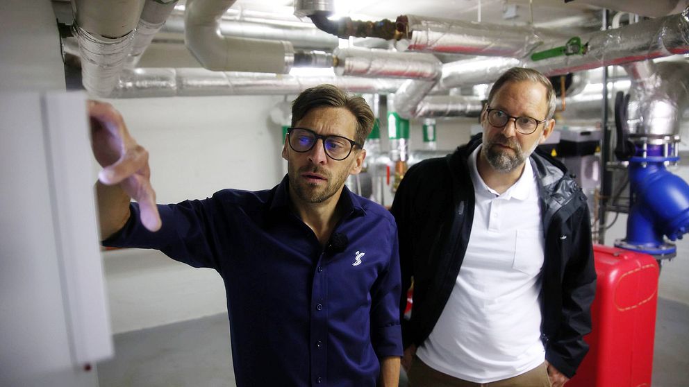 Två män med skägg står i värmecentralen i en källare. Den ena mannen trycker på en skärm som lyser upp hans hand.