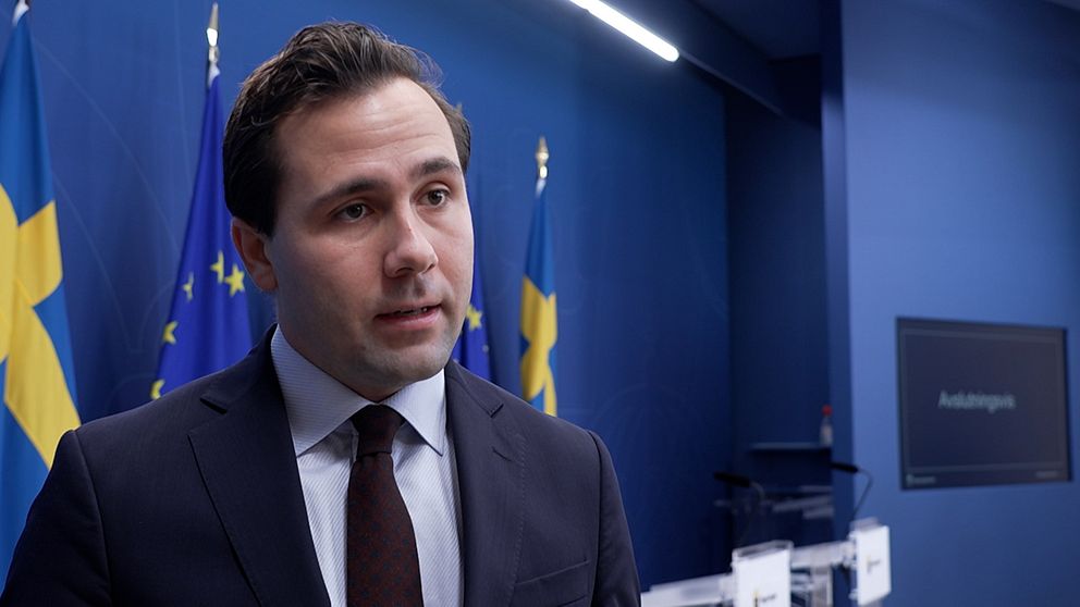 Benjamin Dousa (M) står framför en blå vägg prydd med svenska och europeiska flaggor. I bakgrunden syns en digital skärm.