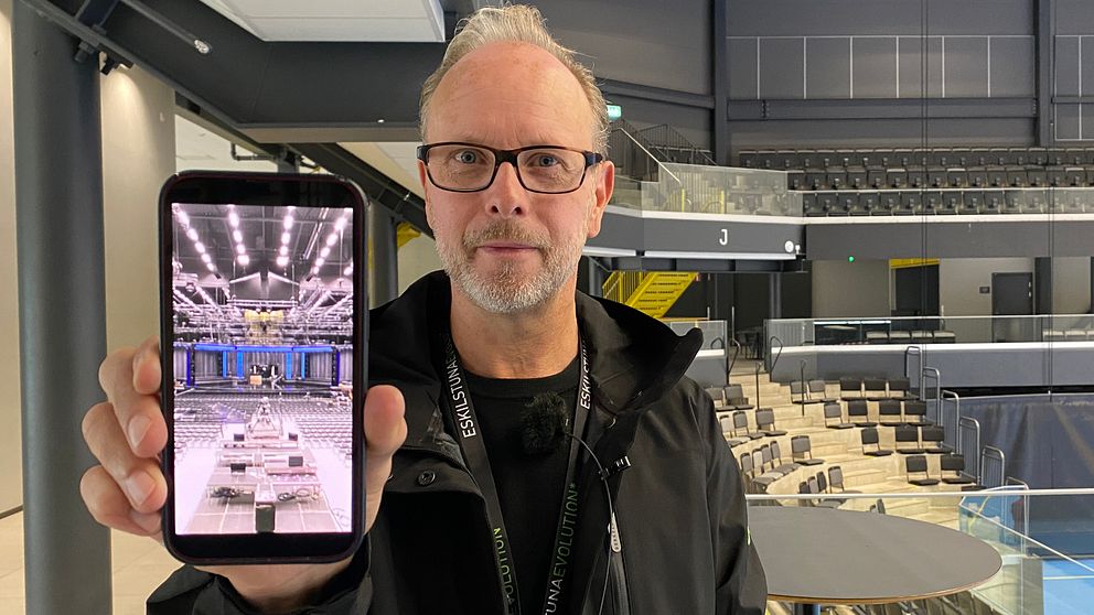 Bild på Micke Lönnberg, evenemangschef på Destination Eskilstuna. Han håller upp en mobiltelefon där han visar en bild på när scenen för Melodifestivalen byggdes i Eskilstuna.