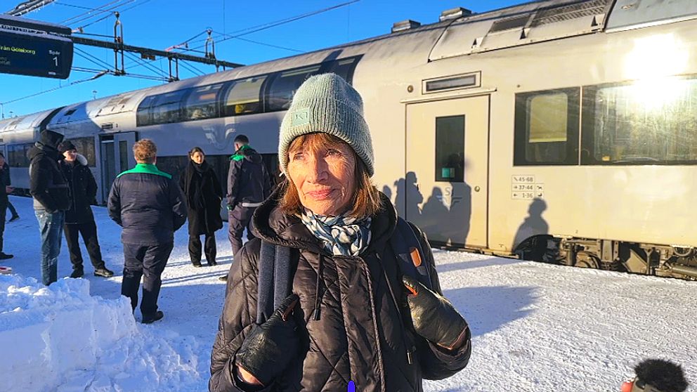 En person står på en snöig tågstation, medan ett tåg är placerat i bakgrunden och flera personer är synliga i närheten. Solen skiner och det finns en digital informationsskylt ovanför.