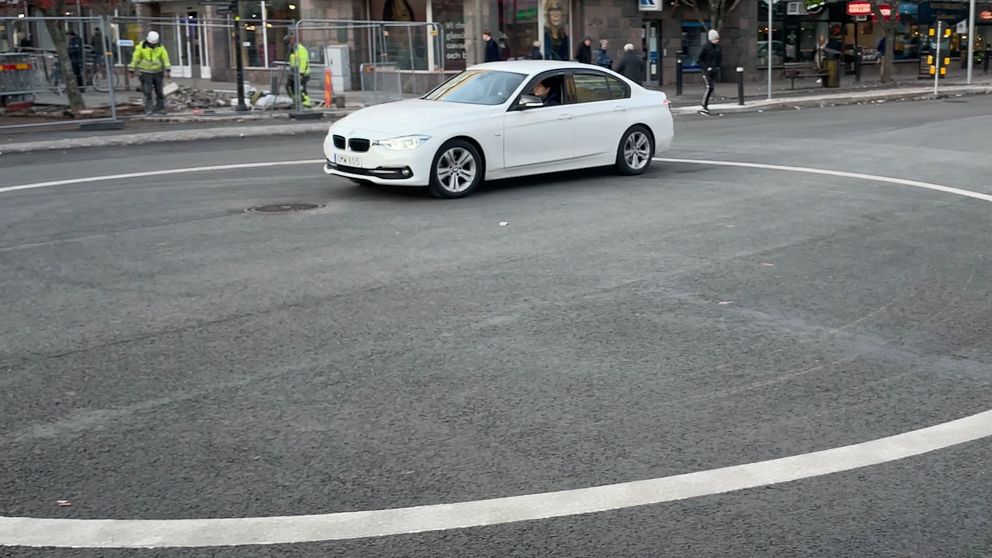 En vit bil svänger i en cirkulerande korsning, med en shoppinggata och byggnader i bakgrunden. Flera personer arbetar i närheten, och en annan bil är synlig längre fram i korsningen.