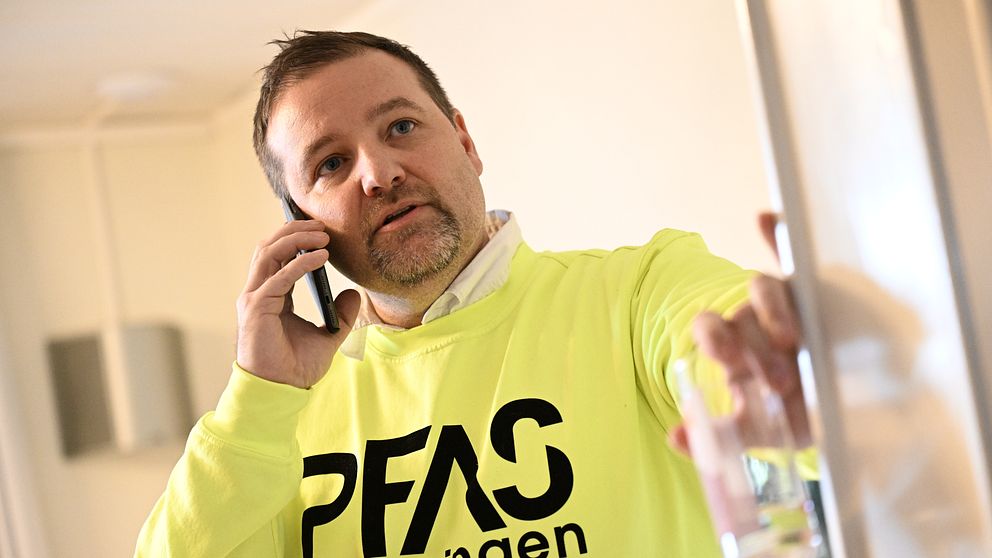 En person iförd en lysande gul tröja med texten ”PFAS-föreningen” står i en inomhusmiljö. Personen pratar i telefon och håller en glasbehållare i ena handen. Bakgrunden är neutral och svagt oskarp, vilket framhäver personen och deras handlingar.