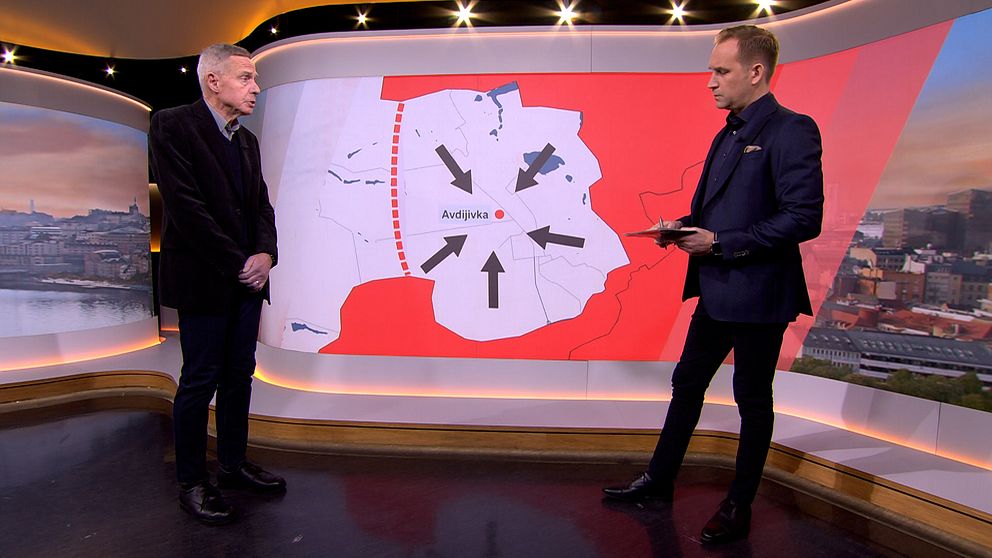 Två män står i studio på varsin sida om en karta med pilar.