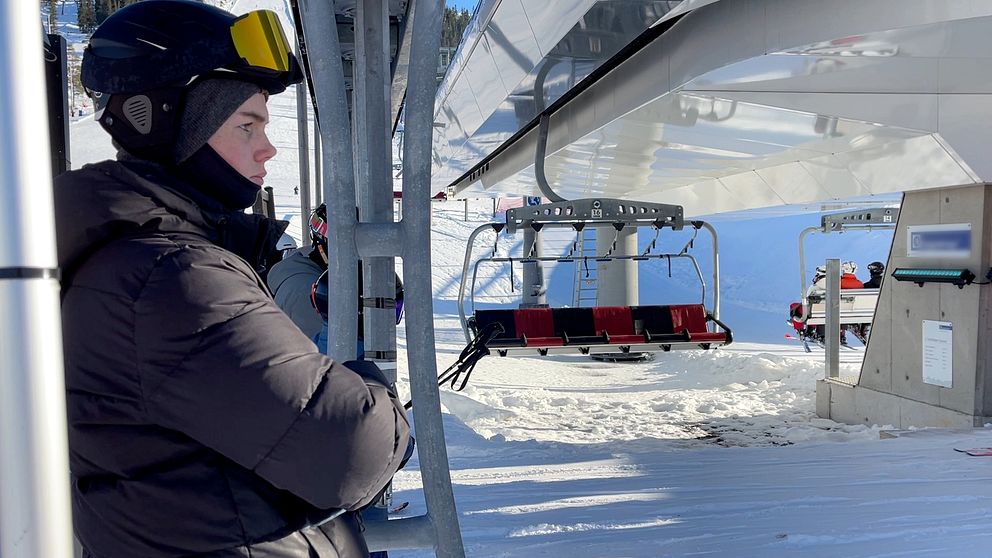 En person iförd hjälm och skidkläder står bredvid en sittlift på en snötäckt skidort, med skidliftstolar i bakgrunden.