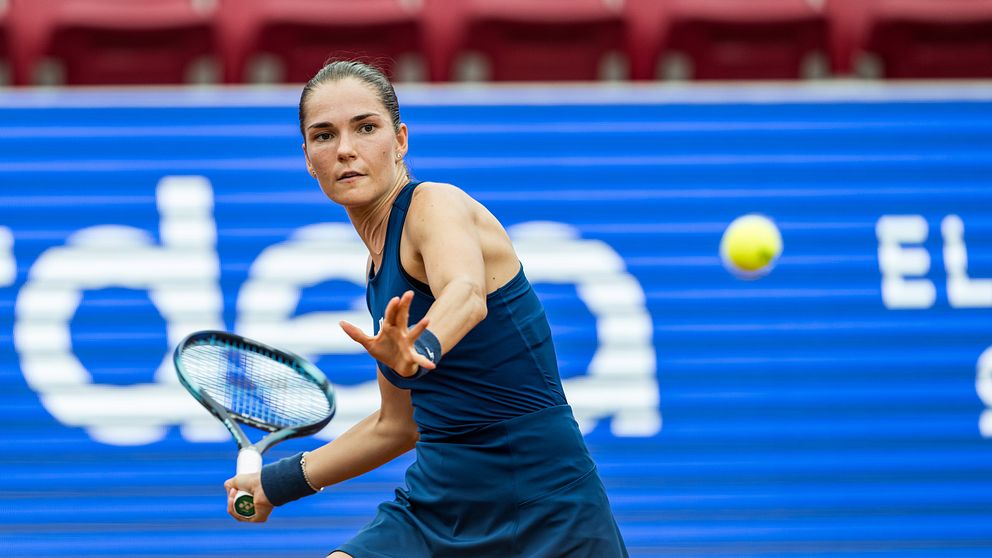 Tennisspelare i blå kläder slår en forehand med racketen på en grusplan, medan en tennisboll är på väg mot dem. Bakgrunden visar en stor reklamskylt med varumärkesnamnet synligt.