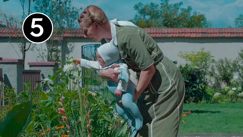 Kvinna håller i en bebis, de böjer sig över en blomrabatt. En scen ur filmen ”Zone of interest”