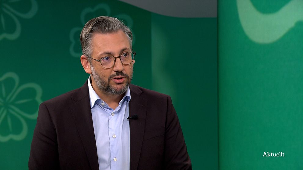 En manlig talare iklädd en mörk kostymjacka och en ljus skjorta står framför en grön bakgrund med blommönster. Bilden verkar vara tagen under en tv-sändning eller intervju. På skärmen syns texten ”Aktuellt.”