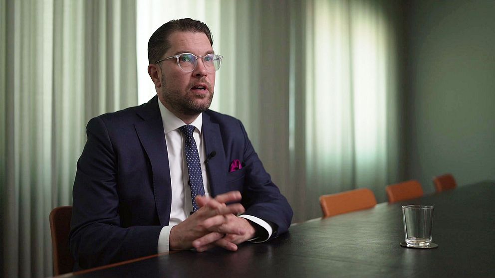 Synkbild på Jimmie Åkesson i intervju med SVT. Åkesson är klädd i kostym och sitter med händerna knäppta vid ett stort bord i ett konferensrum med fördragna garginer i dovt ljus.