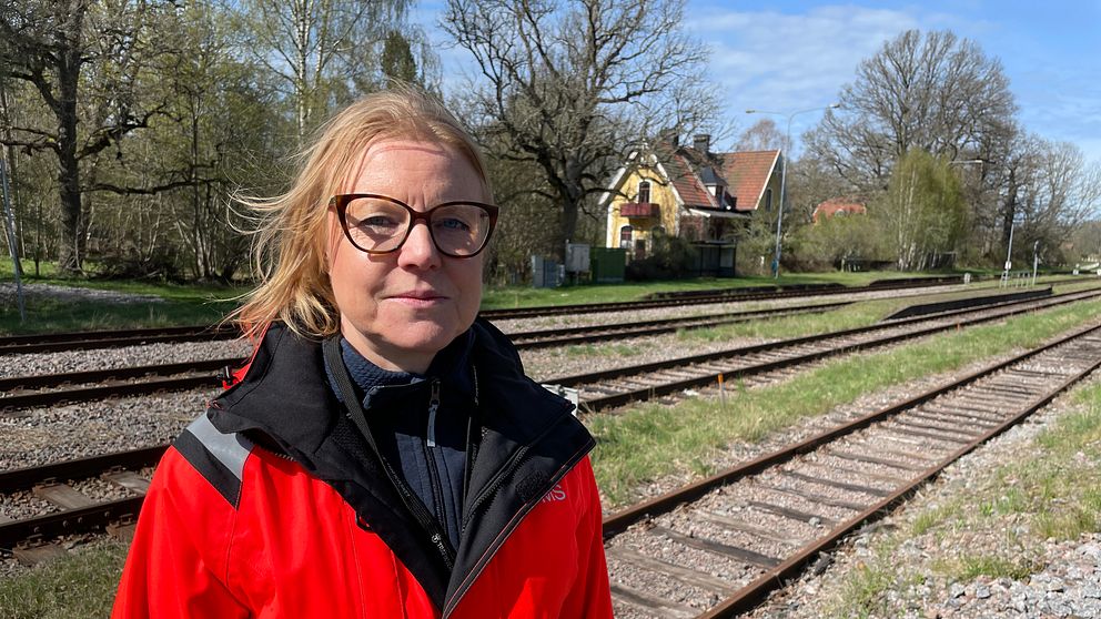 Person i röd reflexjacka står vid ett tågspår med hus och träd i bakgrunden.