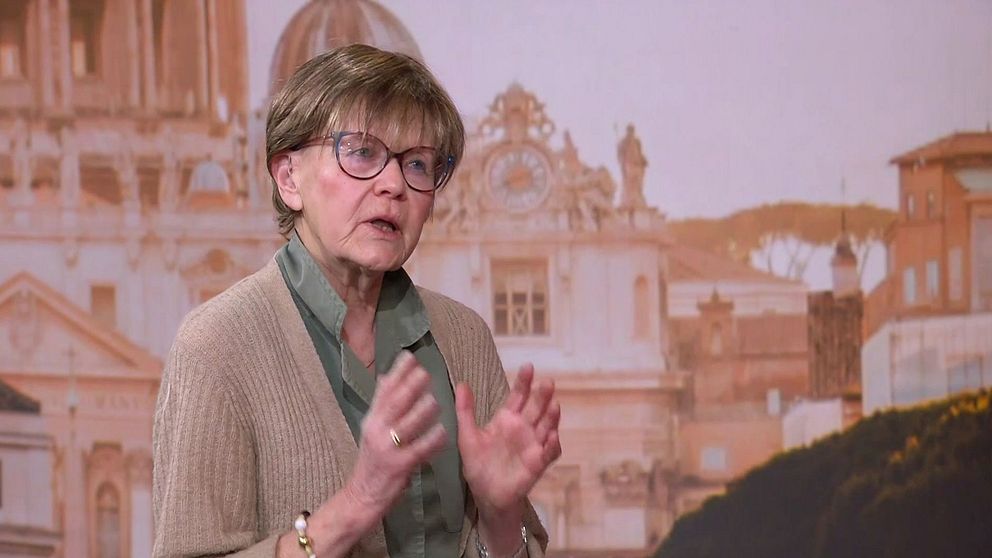 Person gestikulerar framför en bakgrund med klassisk arkitektur och kupolprydda byggnader. I bild syns Vatikankännaren Ulla Gudmundsson.