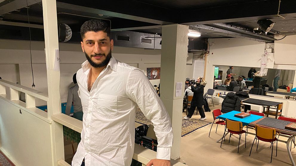 Nadjar Sheikho lutar sig mot en räcke i en lokal i Köping som används som fritidsgård. I bakgrunden syns människor som rör sig runt bord och stolar i olika färger.