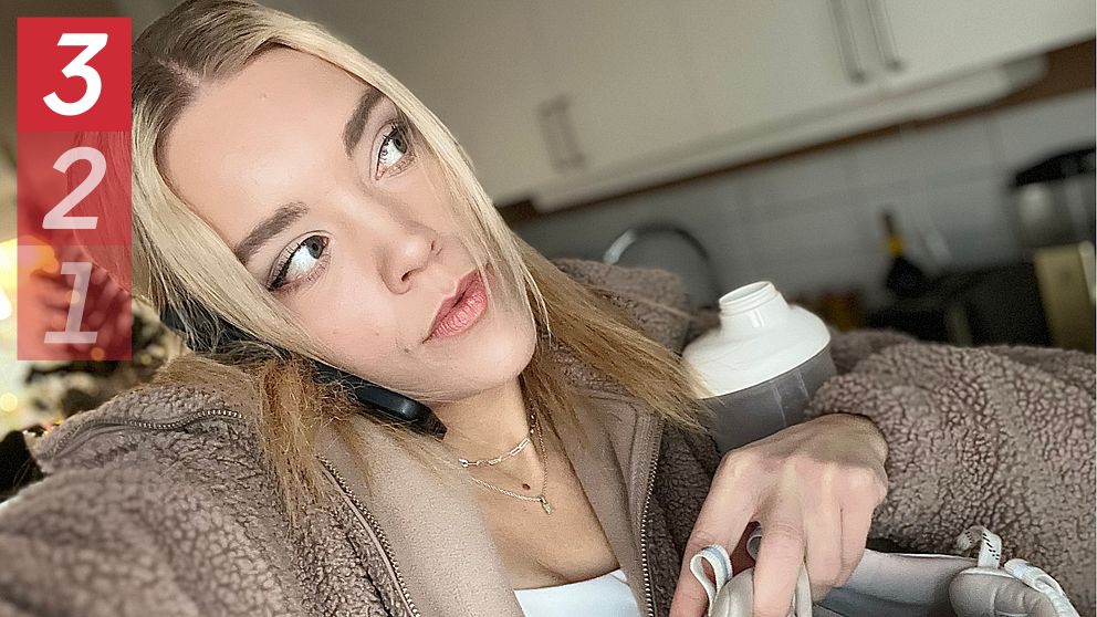 En person i en beige, fluffig jacka håller i en vattenflaska och en sportväska, med en köksmiljö i bakgrunden. Det syns även en julgran i det suddiga bakgrun.