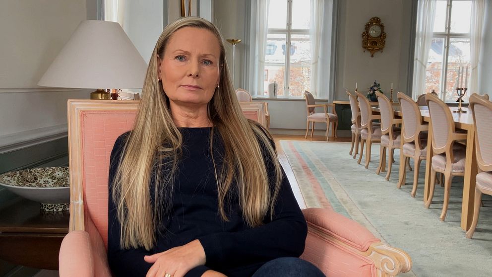 En kvinna i långt ljust hår, Ulrika Gustafsson Lindberg, sitter i en rosa fåtölj i en stor sal.