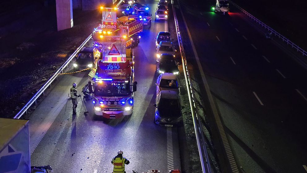 Nattbild av en motorvägsolycka med flera räddningsfordon på plats. Brandmän i reflekterande kläder arbetar vid olycksplatsen, där flera bilar står uppradade längs vägen. Bärgning och säkerhetsåtgärder pågår.