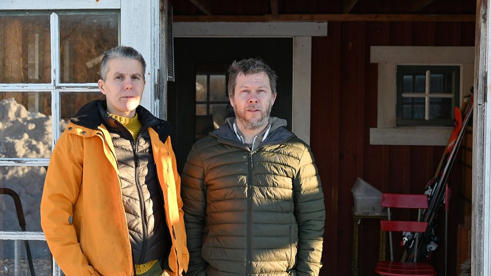 Två personer står vid en stuga med en träveranda, klädda i varma ytterkläder. Den ena har på sig en orange jacka, medan den andra bär en grön jacka. I bakgrunden syns snö och skidor lutade mot väggen.