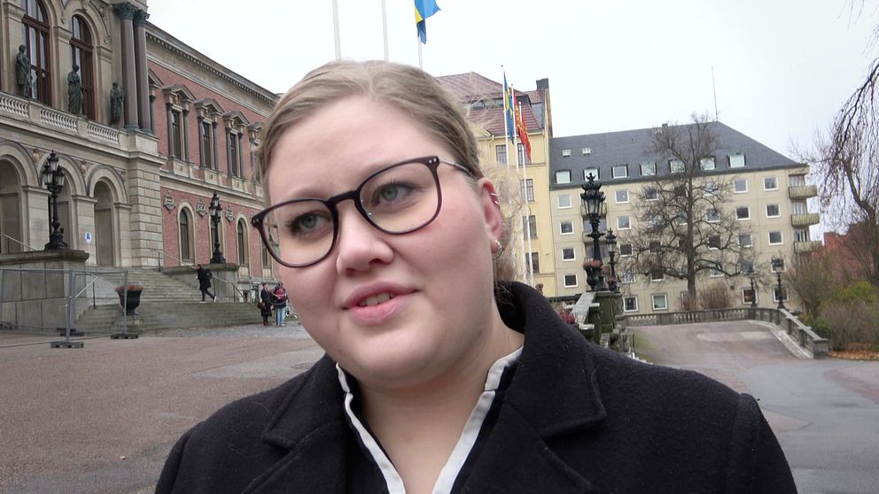 Personen står framför en historisk byggnad med svensk flagga i bakgrunden. Det är en grå och molnig dag, och omgivningen ser ut att vara en stadsmiljö med träd och fler byggnader synliga. Atmosfären verkar lugn och stillsam.