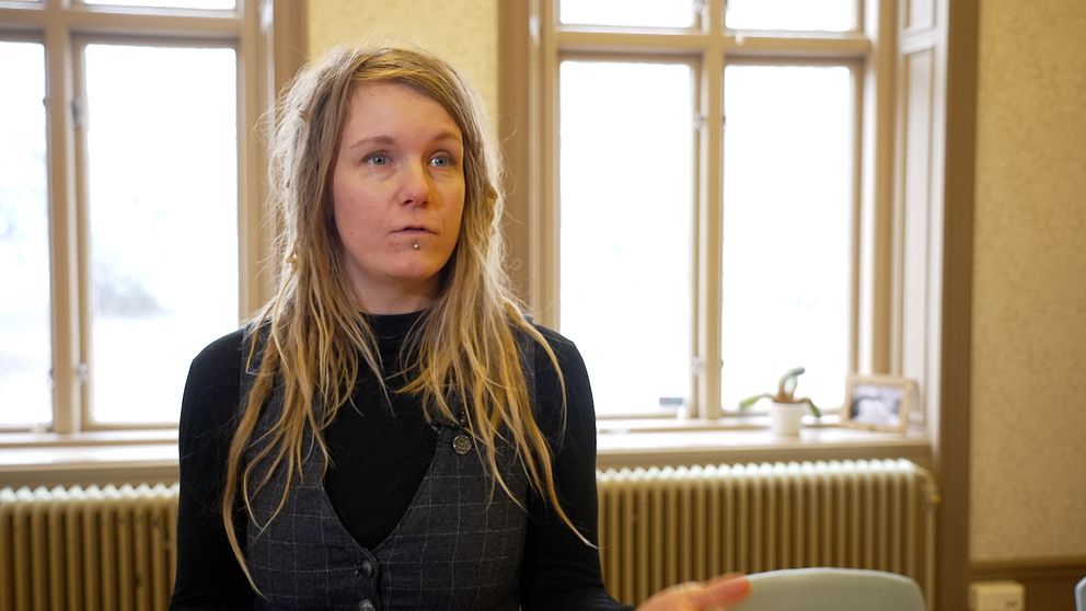 En person med långt blont hår står inomhus framför flera stora fönster och pratar.  Detaljerade inredningselement som en vägg med radiator och möbler är synliga i bakgrunden.