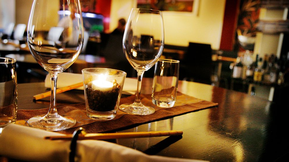 Ett dukat bord i en restaurang med tre vinglas, ett ljus i en glasbehållare med mörka stenar, och en liten shotglas. I bakgrunden syns oskarpa silhuetter av gäster och en bar.