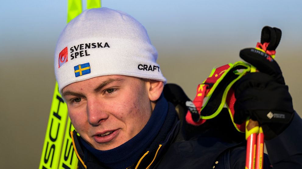 En person står med skidor och stavar, iförd en ljus mössa med texten ”SVENSKA” samt en jacka. Skidorna och stavarna hålls i ena handen, och bakgrunden är suddig men visar en kall och klar miljö.
