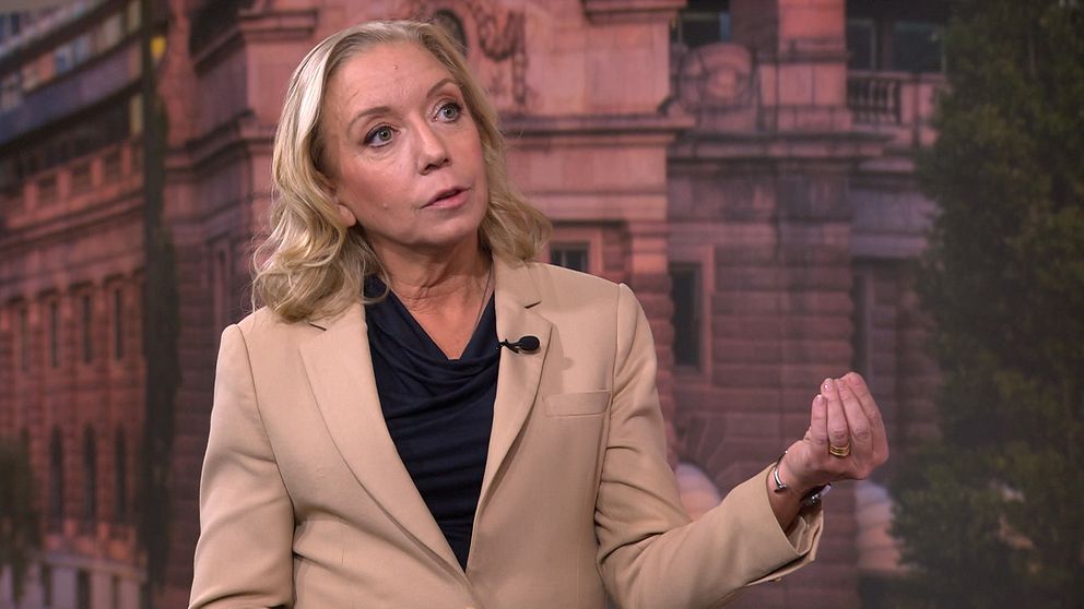 Person i beige kavaj gestikulerar framför byggnad.