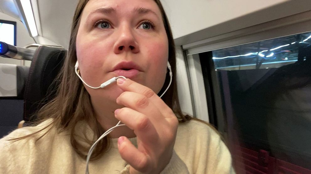 Kvinna i brunt hår sitter i en tågkupé, i tågets tysta avdelning, och håller en hörlur i handen. Bakgrunden visar fönstret i tågkupén, med svagt ljus och en del av järnvägsstationen synlig utanför. Personen har på sig en ljus tröja och ser ut att vara upptagen med att lyssna på något.