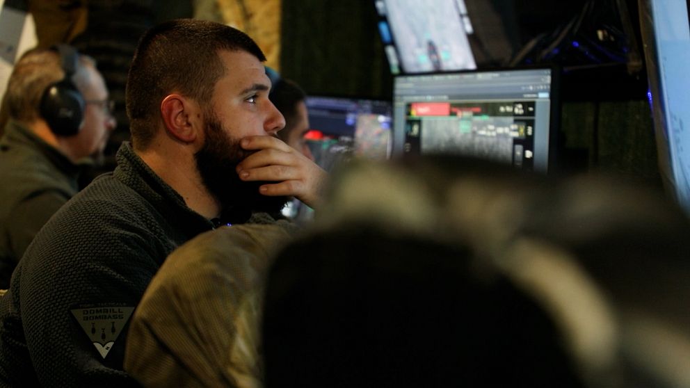 En person sitter vid en dator med flera skärmar, där olika grafiska data visas. Miljön ser ut att vara en kontrollcentral, med fokus på monitorerna och det strategiska arbetet som pågår. I bakgrunden syns andra personer, som också är engagerade i sina uppgifter.
