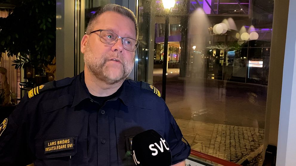 Person i polisuniform med namnskylt ”Lars Broms”, hastighetspolis, som håller en mikrofon med SVT-logotyp. Bakgrunden visar en nattlig stadsmiljö med oskarpa ljus och fönster.