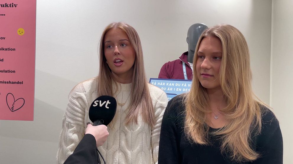 Två personer står framför en vägg med information. . Väggen har olika meddelanden och symboler relaterade till ett ämne, troligen kopplat till utbildning eller information.